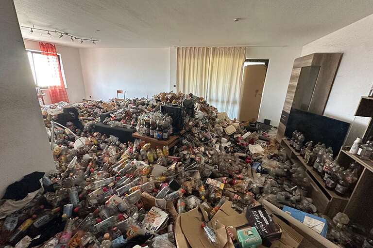 Ein Wohnzimmer, das vollgestopft ist mit Müll, hauptsächlich Plastikflaschen, Lebensmittelbehältern und Pappkartons. Der Müll bedeckt den Boden und verdeckt die Möbel bis auf einen Fernseher und Regale. Notrettung: Messie Wohnung Entrümpelung